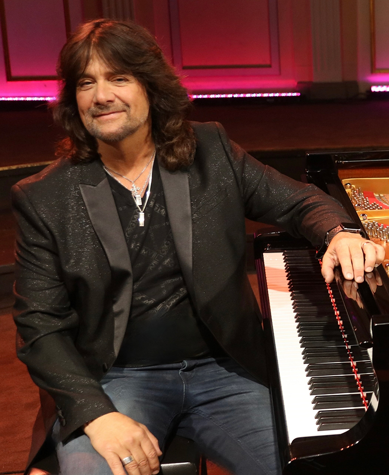 Robert Wells sits smiling with his arm resting on a grand piano